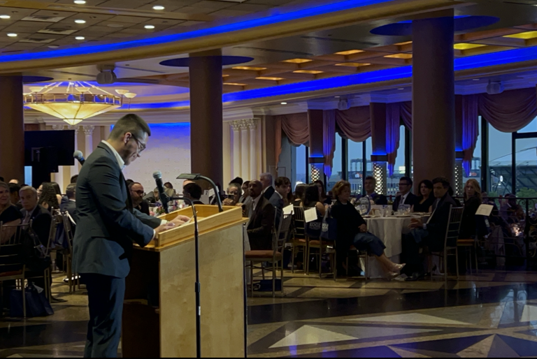 Julio Fernando Salas Vargas stands at a podium in a ballroom. He's wearing a suit and glasses and is smiling as he speaks to people seated at round tables.