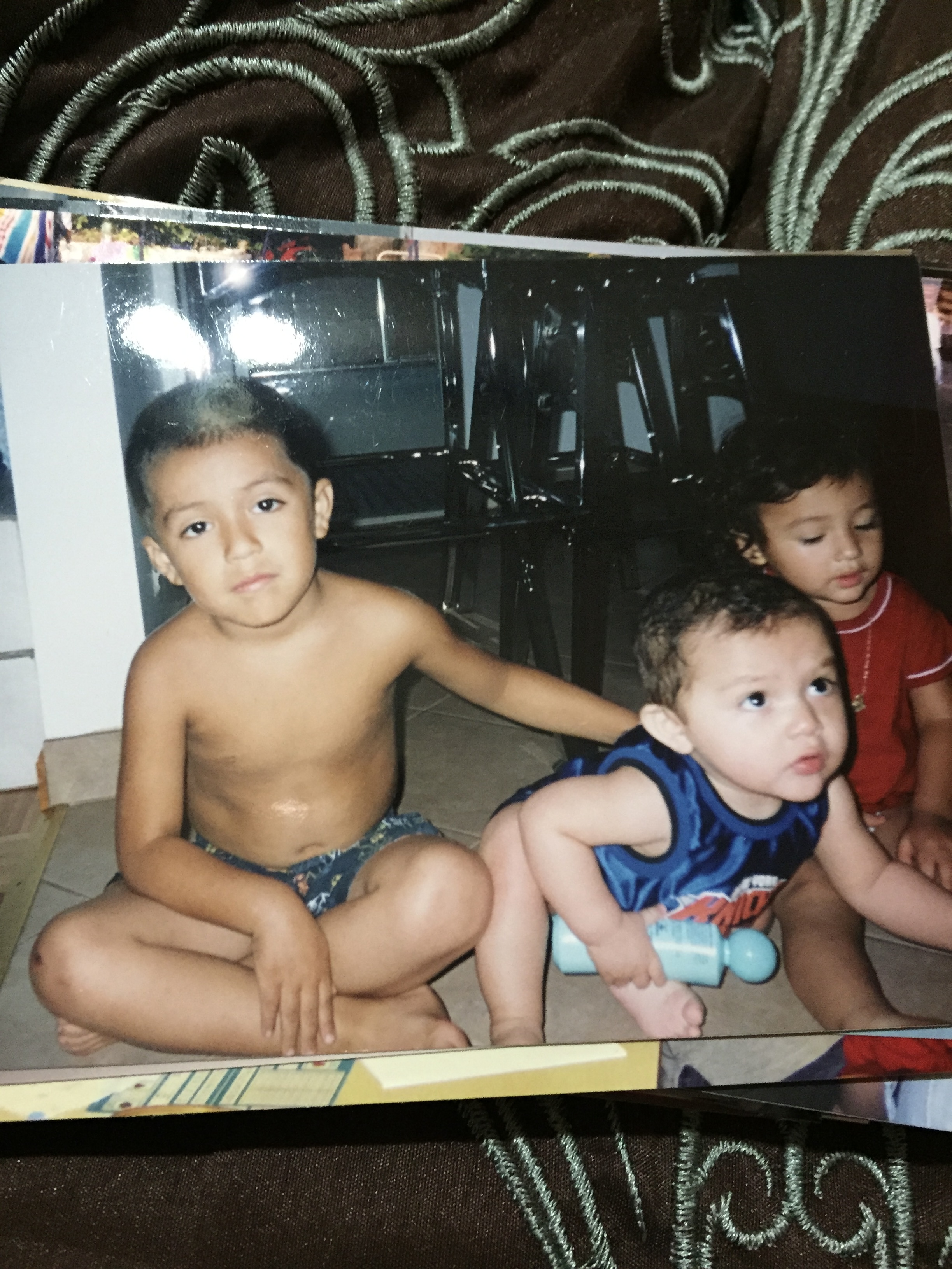 A stack of glossy photos on a brown tablecloth, with the top photo showing three young children with short black hair sitting on a kitchen floor.