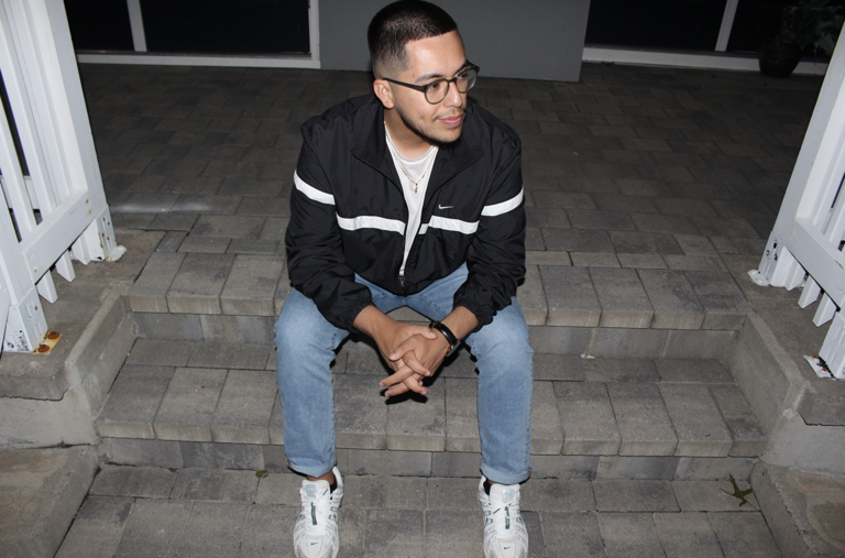 Julio Fernando Salas Vargas leaning forward with his hands clasped, sitting on the gray brick front steps of a building. He has short black hair, a short beard, and wears round black glasses, a black windbreaker, light jeans, and white sneakers.